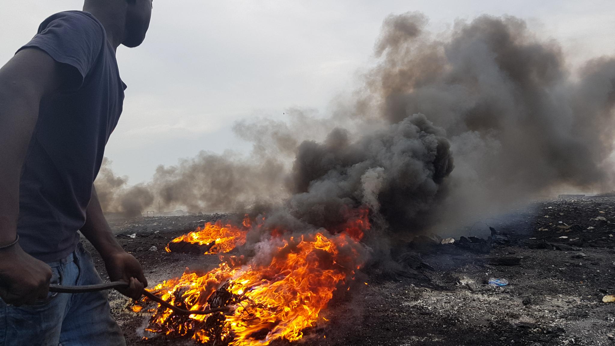 A worker uses a metal hook to tend a fire of burning cables at the Agbogbloshie e-waste site in Accra, with thick black smoke rising behind him.