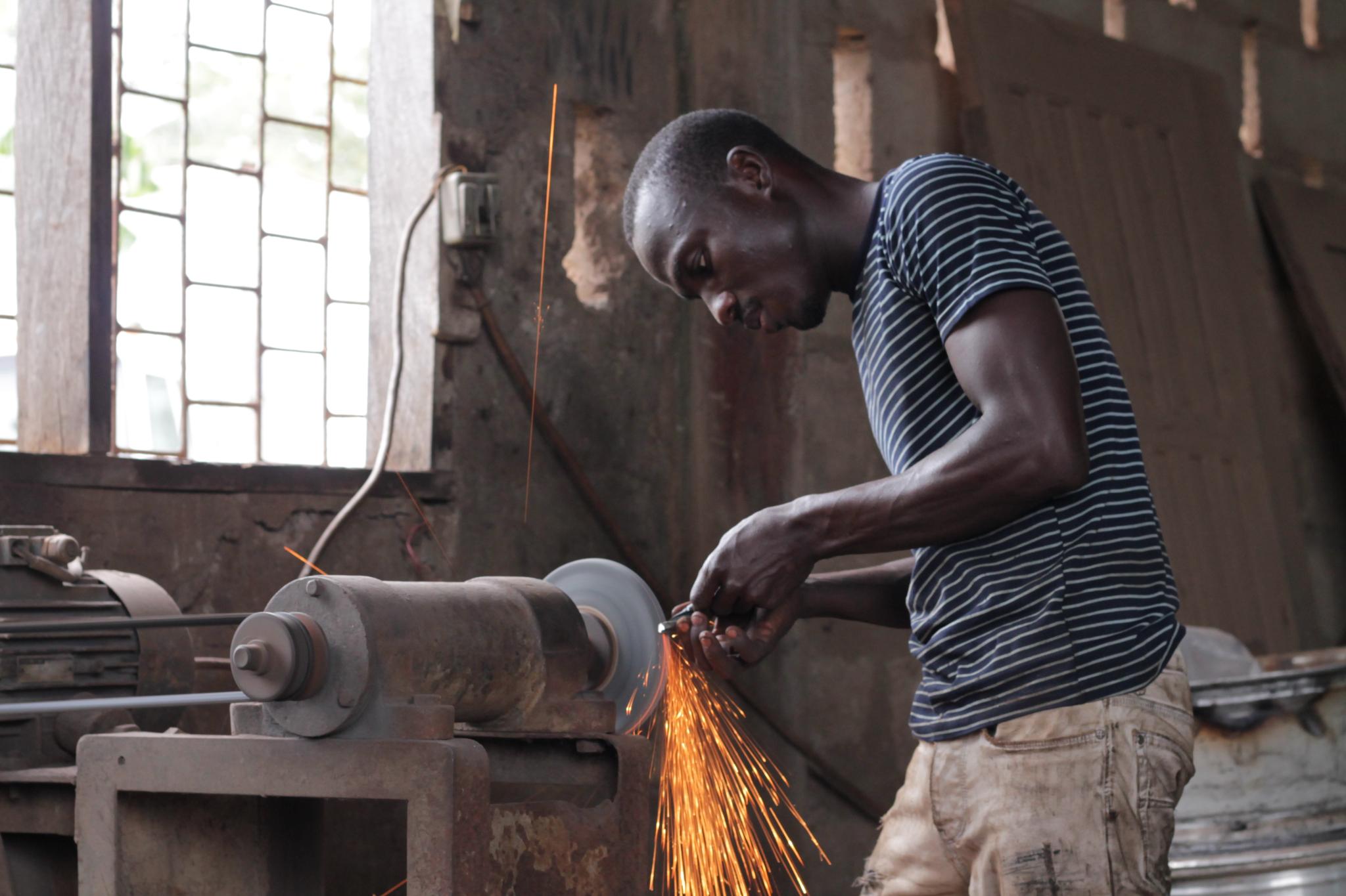 A Ghanaian metalworker grinds a piece of metal on an unguarded bench grinder, sparks flying, without gloves or eye protection.