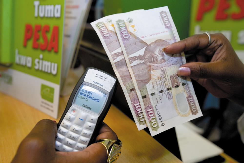 A person holds a Nokia 1100 showing an M-PESA cash transfer prompt while counting Kenyan shilling notes at an agent counter.